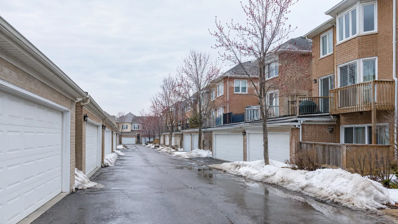 Wet rear lane in a spring thaw, lined with white garage doors and melting snowbanks, with the balconies and bay windows of brick townhouses above