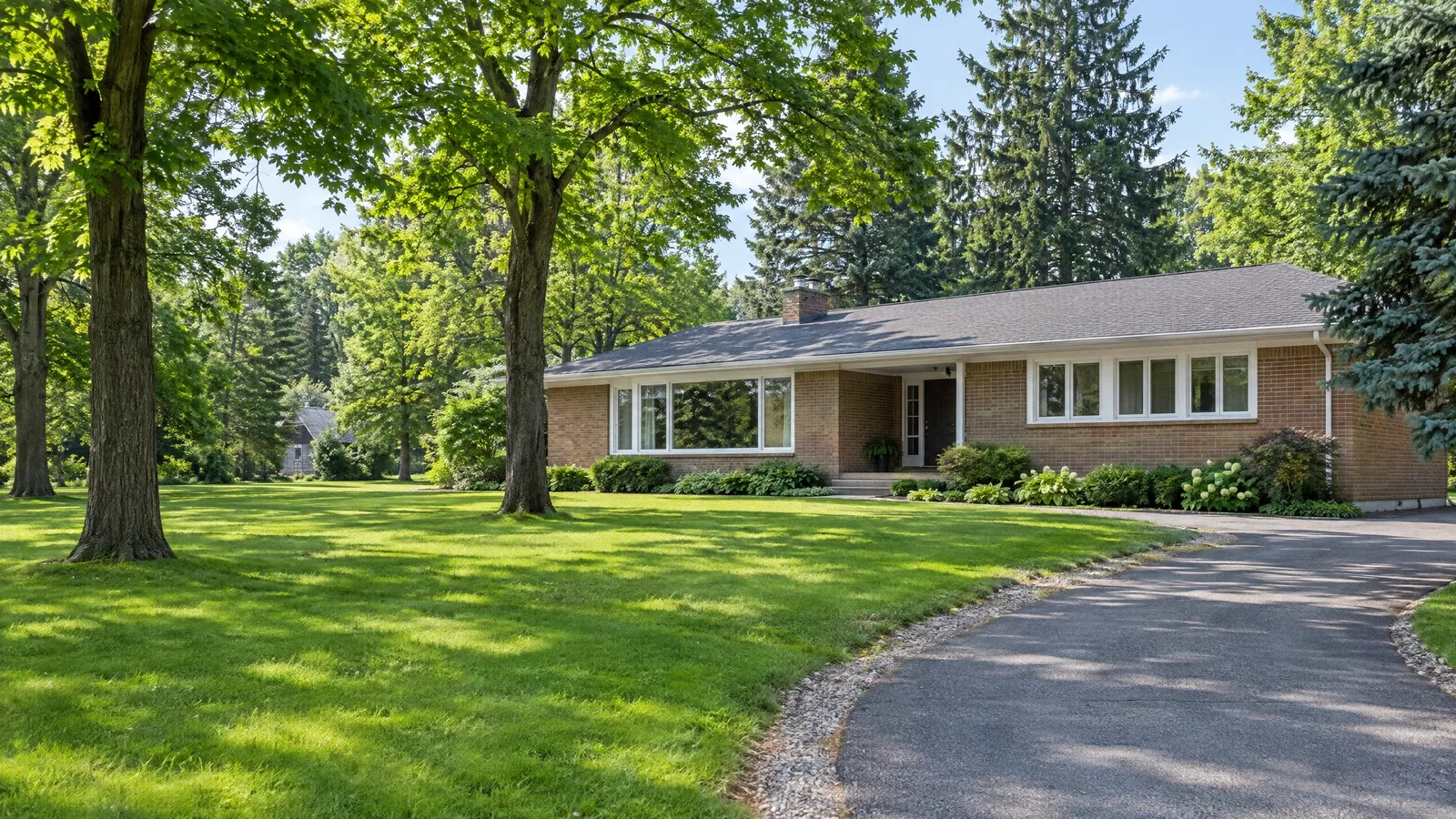 Long red-brick 1960s ranch house with a wide picture window and a row of smaller windows, set back on a broad lawn under mature trees in summer