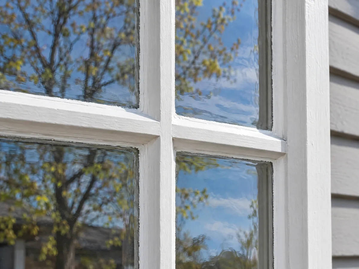 Close-up of a white-painted wood sash where two muntin bars cross, each small pane bedded in glazing putty, the old glass slightly rippled and reflecting blue sky and budding branches