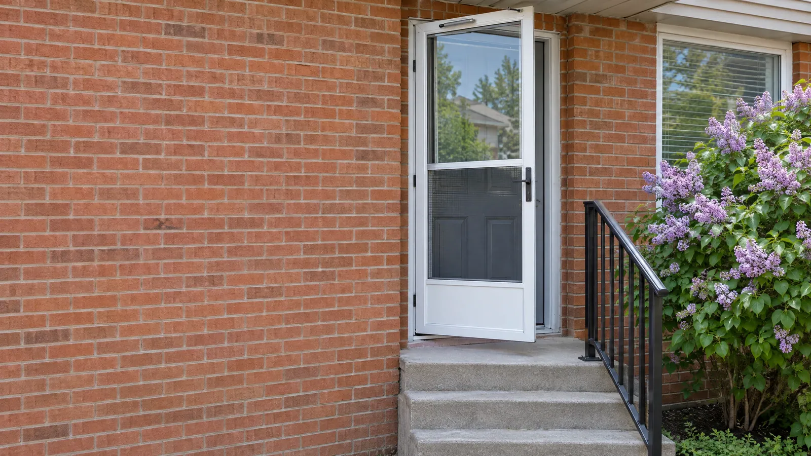 White ventilating storm door propped open at a red-brick side entrance, glass in the upper half and a screen below, beside a black handrail and lilacs in bloom